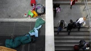 A homeless person sleeps on a metro ventilation grill as commuters enter Syntagma station in Athens