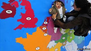 A mother holds a baby over a floor map of Europe