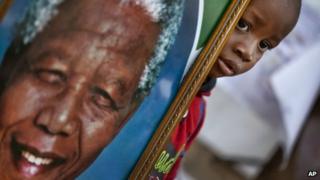 A young boy poses for a photograph of him holding a portrait of Nelson Mandela next to a wall of get-well messages and flowers laid outside the Mediclinic Heart Hospital where former South African President Nelson Mandela is being treated in Pretoria, South Africa Monday, July 8, 2013.