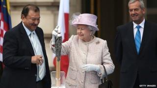 Королева помещает свое послание в замысловатую головку эстафеты, за которой наблюдают президент Федерации Игр Содружества Принц Имран Тунку и лорд Смит из Кельвина, который возглавляет Оргкомитет Глазго 2014.