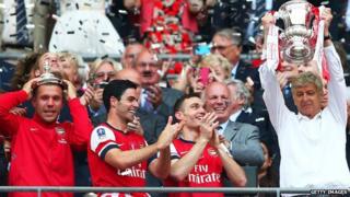 Arsene Wenger lifts a trophy in celebration alongside Lukas Podolski, Mikel Arteta and Thomas Vermaelen