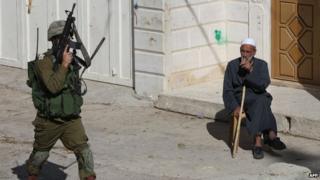 An Israeli soldier walks past a Palestinian man sitting on a street in the West Bank town of Hebron