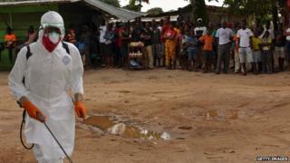 A public health worker in Liberia disinfects a courtyard as villagers watch.