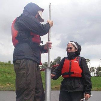 Two scientists stand on either side of a tall metal cylinder. They are on a platform in a lake.