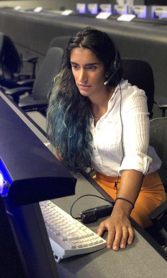 A woman with long brown and green hair, a white top and yellow skirt sits at a desk in JPL's mission control, wearing a communications headset