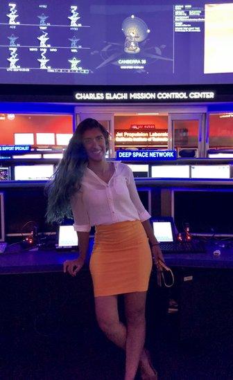 A woman stands in front of signs saying 'Charles Elachi Mission Control Center' and 'Deep Space Network'. Above are screens with radio telescopes and the data they are receiving