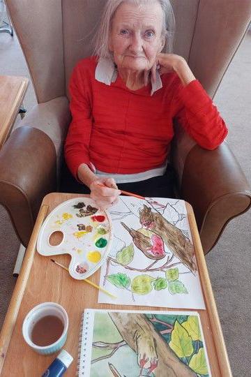 An elderly woman in a red jumper paints a picture of a robin