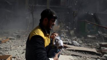 Amid rubble and smoke, a man wearing a headlamp and yellow jacket carries a baby in distress