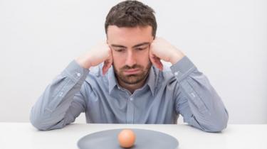 Hombre joven mirando a un plato con un huevo con cáscara.