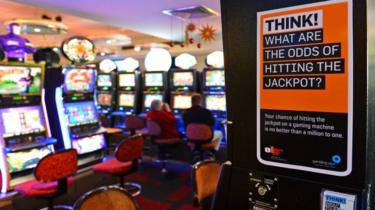 A room with poker machines, or slot machines, behind a warning poster saying: "Think! What are the odds of hitting the jackpot?"