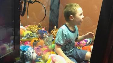 A young boy sits inside an arcade game machine on top of stuffed toys.