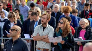 A crowd of people watch the ceremony, some with heads bowed, some reading from service sheets, in Hagley Park on March 29