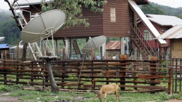 Une chèvre mange de l'herbe près des antennes paraboliques installées à l'extérieur des maisons d'un village du plateau de Nakai au sud-ouest du Laos.