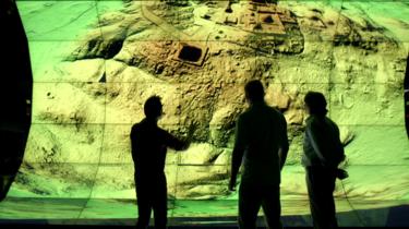 Trois géomètres regardent un grand écran numérique montrant une image Lidar d'une ville maya.