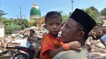 An Indonesian man holds a boy across from collapsed buildings as search and rescue members search for a victim of the recent quake in Tanjung on Lombok island on August 9, 2018.