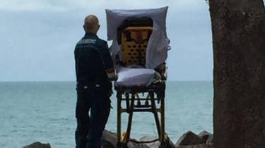 A paramedic stands behind a wheeled stretcher carrying a terminally ill woman, with both looking out at a beach