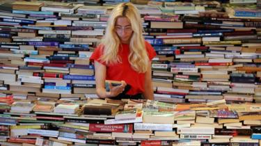 blondýnka stojí uprostřed obrovské hromady knih.