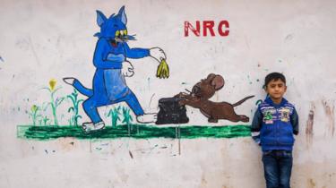 Een kind in het Syrische vluchtelingenkamp Domiz in 2014 staat naast een muurschildering van Tom en Jerry. NRC staat voor Norwegian Refugee Council