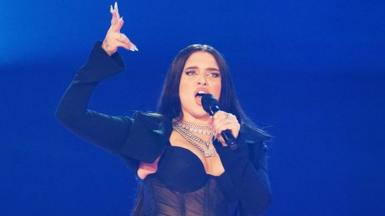 Mae Muller performs on stage at Eurovision. She's wearing a blazer, sheer corset and several chunky silver necklaces. Her hair is down and she's pointing a gun finger in the air.