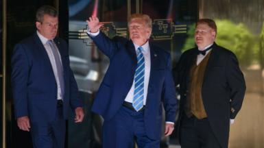Trump in blue suit with blue tie in front of glass doors, between man in blue suit and door man in black suit with gold vest.