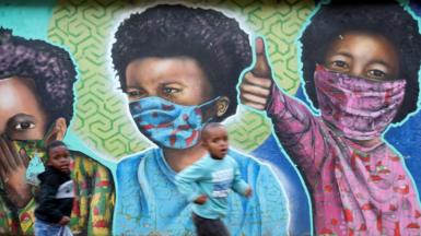Boys run past a bright mural of children wearing face masks.