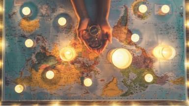 Hands hold a compass over a world map, with candles across it