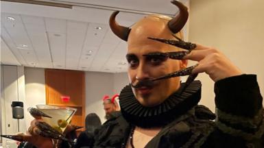 A man wearing horns, a black ruff and elaborate pointed fake nails poses with a martini