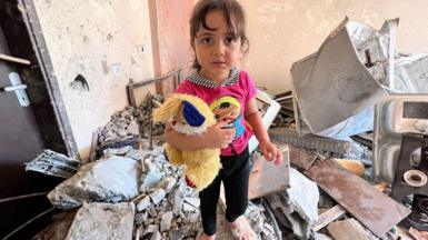 Tuta standing on rubble of a destroyed home carrying a yellow teddy bear