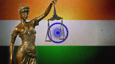 A small bronze statuette of Lady Justice before an Indian flag.