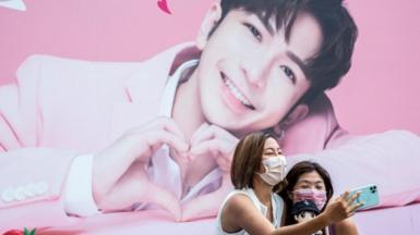 This picture taken on August 6, 2021 shows two women taking a selfie in front of a billboard showing a portrait of Anson Lo, a member of Cantopop boyband Mirror,