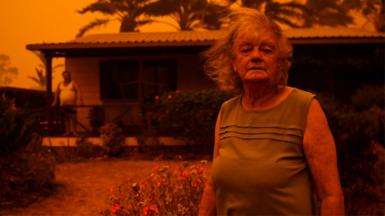 Woman stands in front of home surounded by orange haze from fires