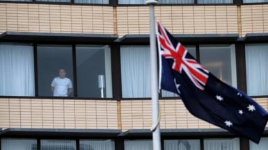 An Australian flag flies in front of a hotel building where a man looks out from his quarantine room