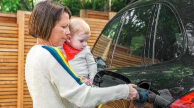 A woman holds a baby whilst charging her electric car