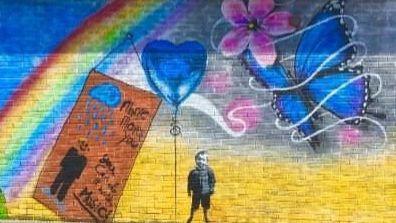 mural on a brick wall featuring a rainbow, a blue butterfly and a small black and white boy
