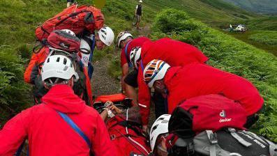 Wasdale Mountain Rescue Team carrying a casualty to the waiting Great North Air Ambulance. They are dressed in red and wear helmets.