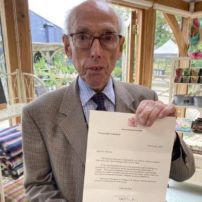 A man in a jacket, shirt and tie holds up a letter addressed to him from the King, on cream paper with a red royal logo at the top, while standing in a gift shop