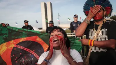 Dois indígenas protestando ao lado de bandeira do Brasil com aparência ensanguentada e prédios do Congresso Nacional atrás
