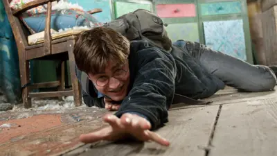 Scene from film with Daniel Radcliffe playing the role of Harry Potter, on a wooden floor reaching out with his left hand. He is wearing round spectacles, a dark jacket and jeans, with a backpack. There is a brown wooden chair behind him with green cabinets in the background.