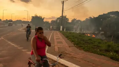 Um homem e uma mulher de bicicleta na estrada passam perto de área verde queimada ao lado; a mulher tapa o nariz
