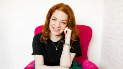 Sara Gibbs sitting in a pink chair, smiling to camera