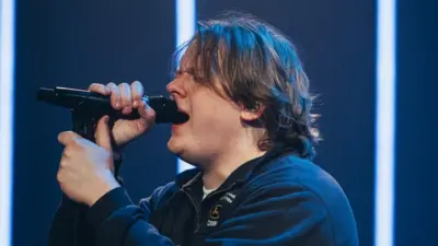 Side profile of Lewis Capaldi singing on stage, holding a microphone close to his mouth. He's in a dark blue shirt and the stage background is the same colour with thin white fluorescent stripes