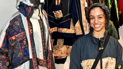 Kazna is smiling wearing a dark head covering and a blue jacket with a light gold and brown print across it. A black bag strap is going across. The background Kazna's designs, one is printed dark blue with white outline. The other on is dark with a printed pattern running across the middle.