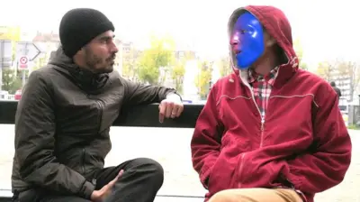 Ahmed Shihab-Eldin entrevistando pessoa com máscara (não identificada), ambos sentados em um banco ao ar livre