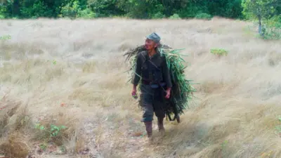 Um homem japonês com uniforme militar caminha em um prado com um disfarce de grama nas costas