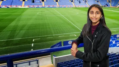 Renuka at Leicester City's stadium. Behind her is the green pitch and empty stands with blue seats, with white writing spelling out Leicester City. Renuka is looking at the camera, leaning on blue railings, with her left hand over her right hand. She is wearing a black jacket with a maroon turtle neck top.