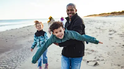 Un padre juega al avión con su hijo en una playa, mientras la hija observa