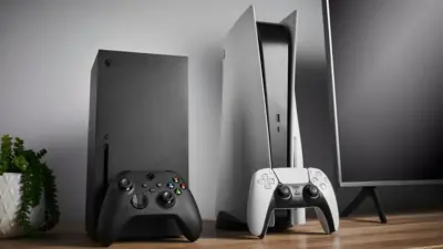An Xbox Series X and Playstation 5 side-by-side on an office desk that's pristine and clean. Both machines are standing vertically, with their respective controllers propped against them. A computer monitor is partially visible on one side of the image and a small potted plant on the other.