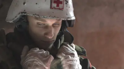Denys wearing a white helmet with a red cross on it. The atmosphere is sombre and he's looking pensive.