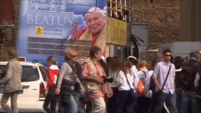 Bus with commemorative beatification decor in back, surrounded by people in Rome