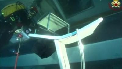 A white chair floating underwater in a submerged part of the Costa Concordia, with a masked diver in the background.